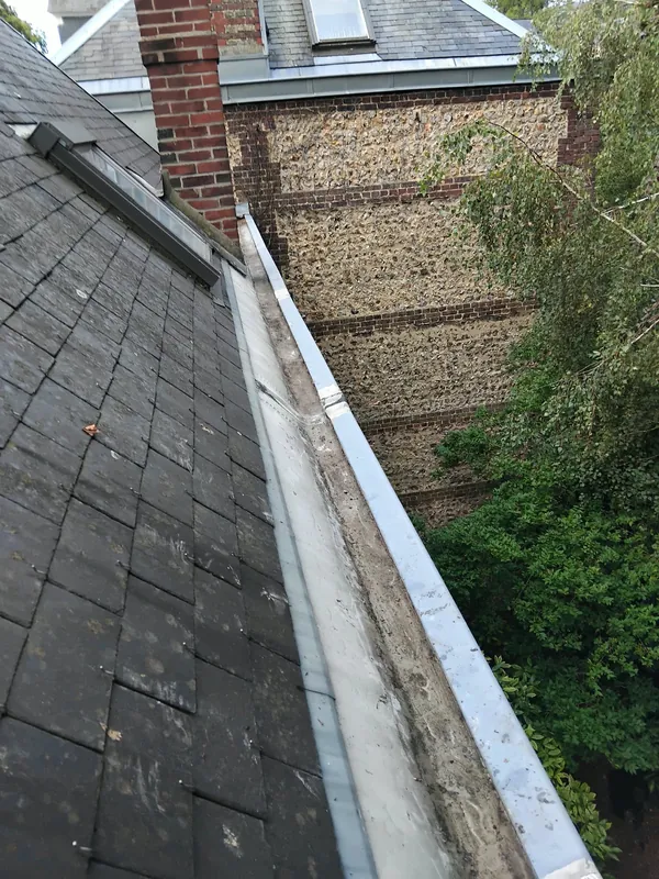 Chéneau en zinc au bas d'une toiture en ardoise, contre un mur en silex et brique, à Franqueville-Saint-Pierre
